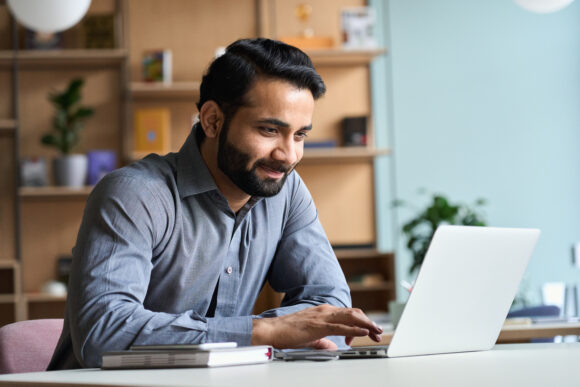 Smiling indian business man working on laptop at home office. Young indian student or remote teacher using computer remote studying, virtual training, watching online education webinar at home office.