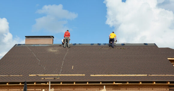 Roofing contractors with fall protection, roof safety harness, ropes are finishing to install asphalt roof shingles on the gable house roof. Roofing construction.