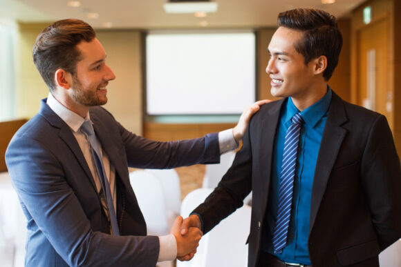 Closeup of two smiling middle-aged business men shaking hands and greeting each other in conference room with whiteboard in background