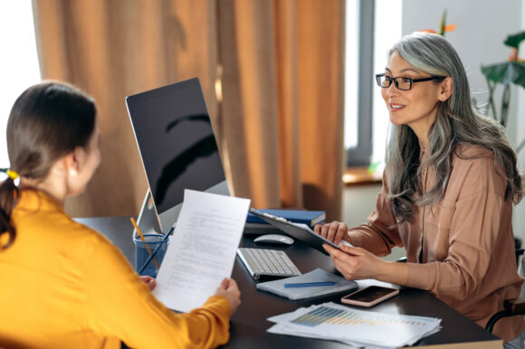 A Mature gray-haired asian woman, employer, examines the resume of a female job seeker, sitting at the workplace. Job interview, hiring, getting a dream job