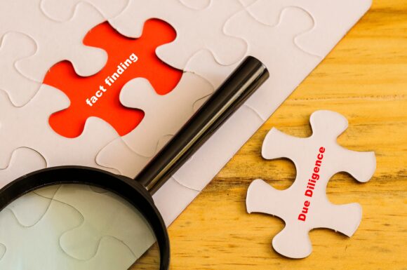 conceptual of due diligence fact finding. Text Due Diligence on loose jigsaw piece. Fact finding on red in place of missing piece. Magnifier on off-white jigsaw puzzle. Selective focus wooden surface.