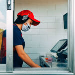 Bangkok, Thailand - May 28, 2020 : fast food cashier in drive thru service waring hygiene face mask to protect coronavirus pandemic or covid-19 virus outbreak working on counter at the station.