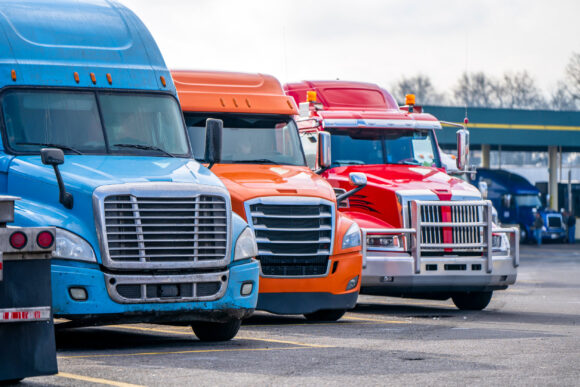 Different bonnet makes and models of professional big rigs semi trucks with commercial cargo on semi trailers standing in row on the industrial truck stop parking lot waiting for unloading