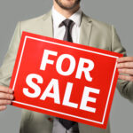 Real estate agent holding "For sale" sign on grey background
