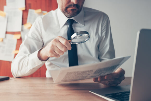 Businessman reading document contract papers with magnifying glass at office desk, close up with selective focus