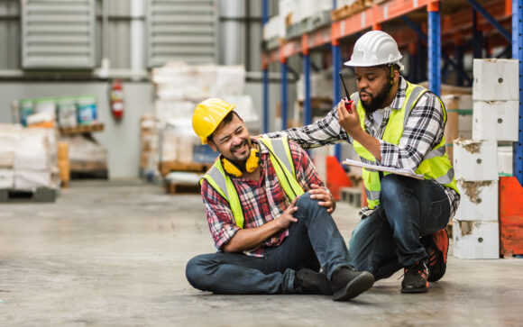 Professional male worker help his colleague after getting accident, injured leg while working in factory or warehouse. Safety, Industry, Healthcare, Insurance Concept