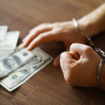Handcuffed Hands Touching Dollar Bills on Wooden Table - Financial Crime Concept