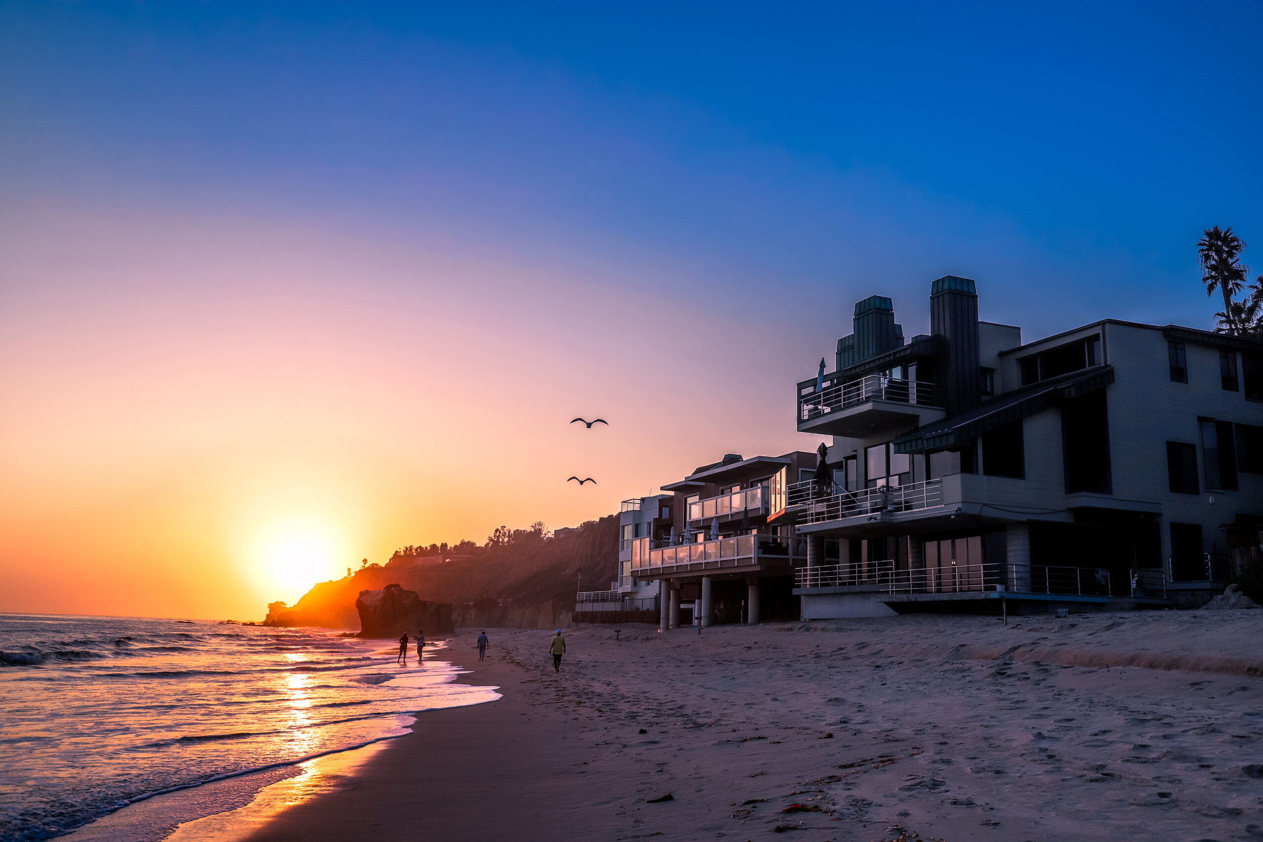 a stunning shot of the sunset at the beach with birds in flight in the sky, homes on the beach and people walking in the sand at El Matador beach in Malibu California USA