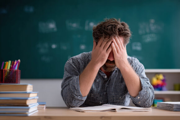 Overwhelmed teacher crying while covering his face with hands, feeling the weight of mistakes and stress in a busy classroom setting