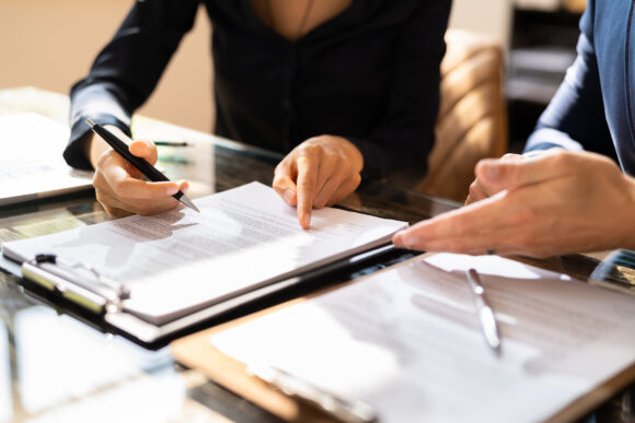 Close-up Of Two Businesspeople Hand Working On Contract Paper Over Desk