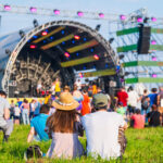 Back view of unrecognizable couple in casual clothes sitting on ground in front of crowd and stage during music festival on sunny summer day