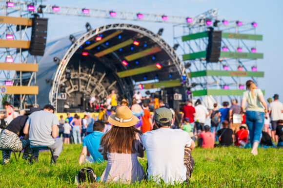 Back view of unrecognizable couple in casual clothes sitting on ground in front of crowd and stage during music festival on sunny summer day