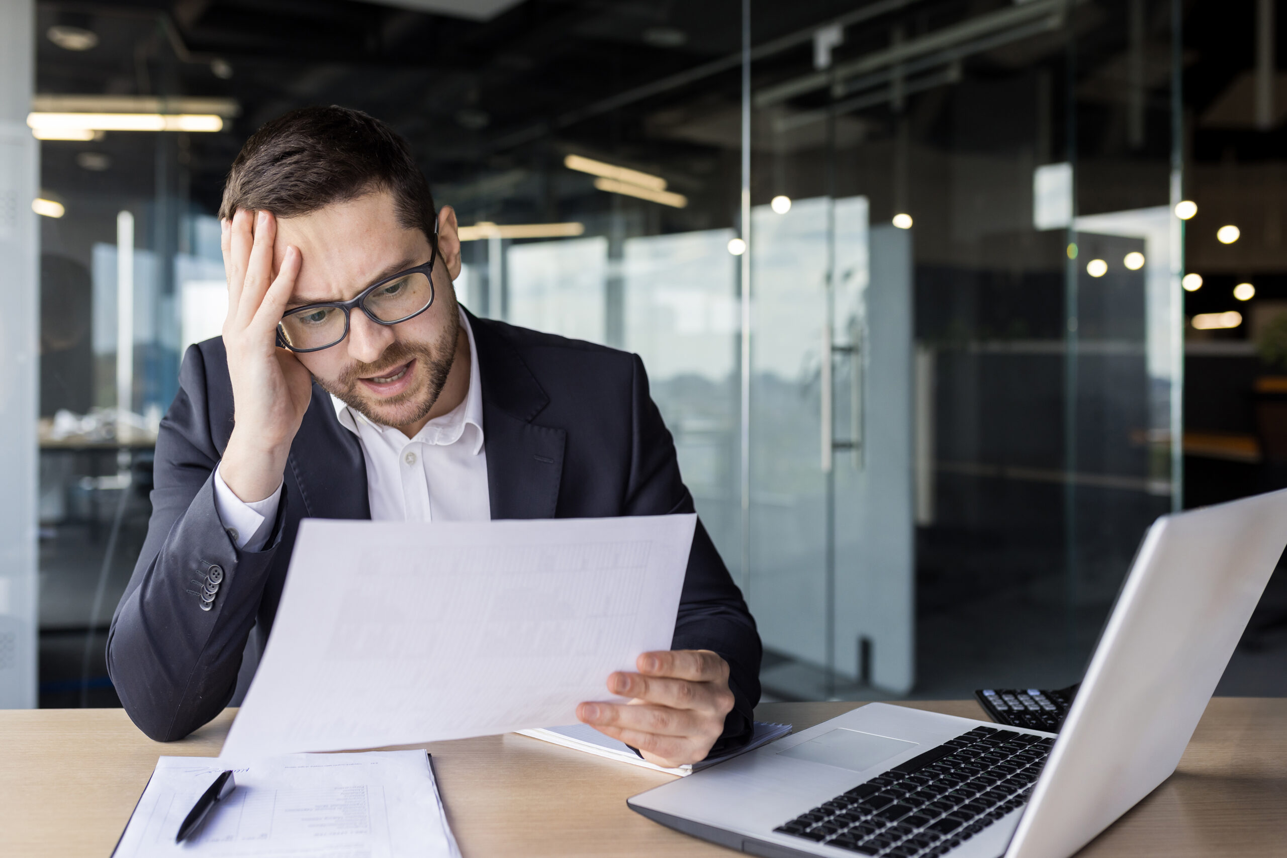 Worried young man businessman, accountant, financier works in the office, holds his head with his hand, looks sadly at documents, bills