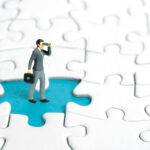 Miniature tiny people toy figure photography. Businessmen standing above missing piece of puzzle jigsaw using binoculars.