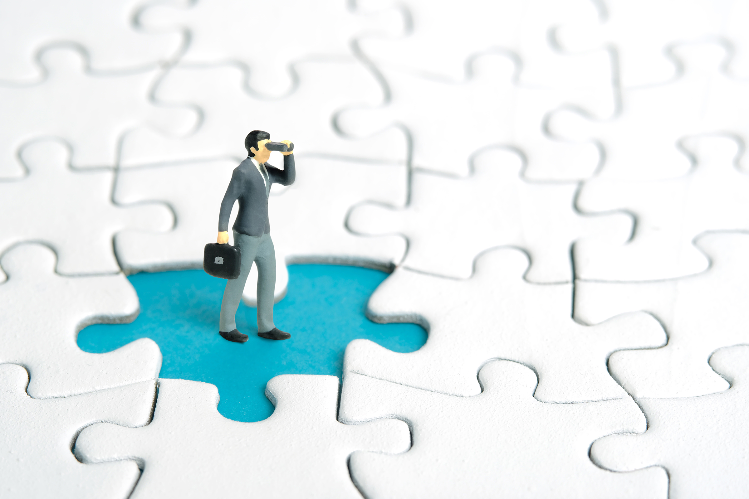Miniature tiny people toy figure photography. Businessmen standing above missing piece of puzzle jigsaw using binoculars.