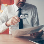 Businessman reading document contract papers with magnifying glass at office desk, close up with selective focus