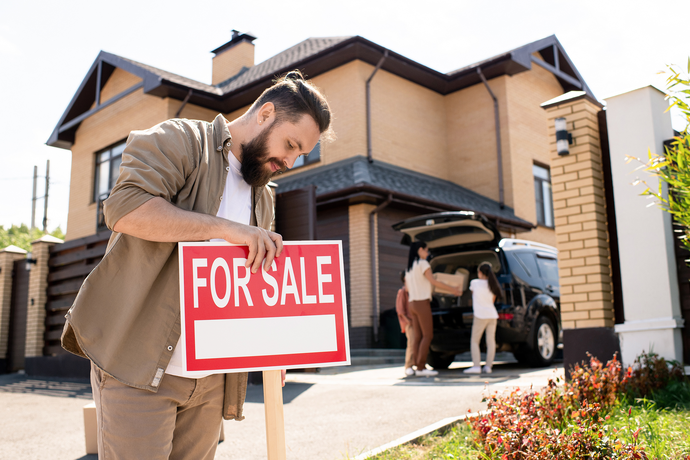 Serious bearded man in casual shirt placing For sale sign against house to pay attention of buyers