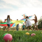 Group of children and teachers playing with rainbow playground parachute on green grass, low angle view. Summer camp activity