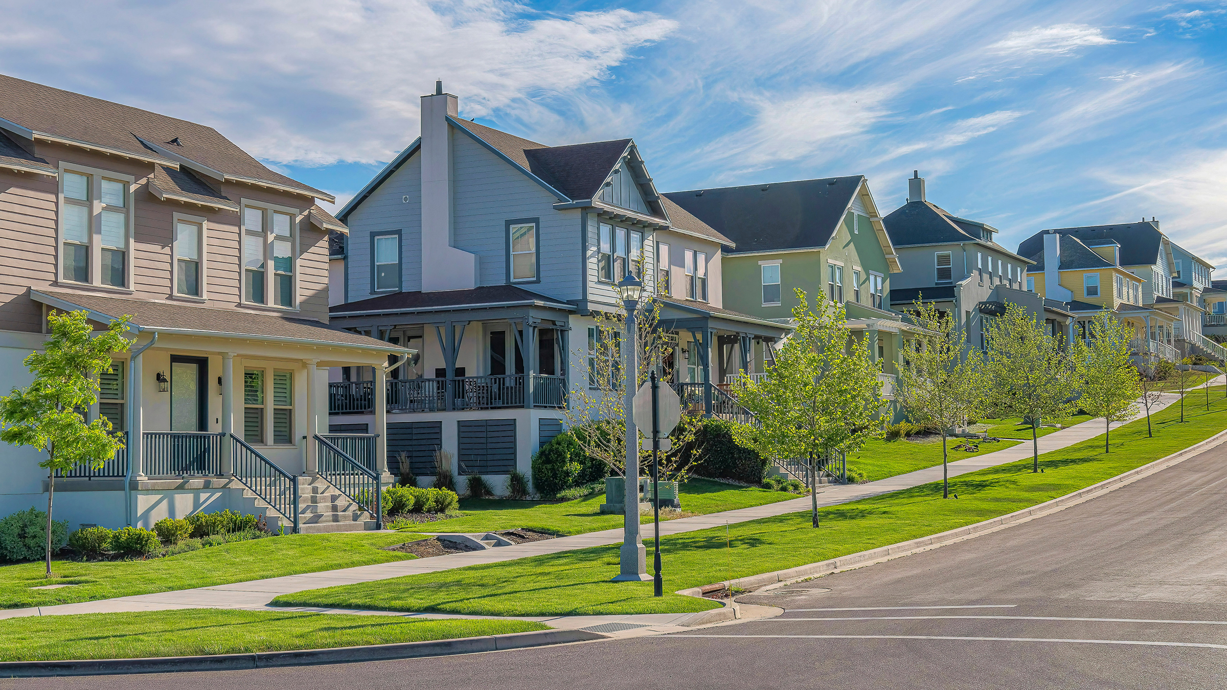 Panorama Whispy white clouds Row of large two-storey houses near the paved uphill road at Daybreak, Utah. View of houses from the intersection road with grass and trees near the sidewalk against the sky and mountains.