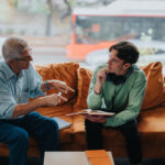 An elderly mentor providing guidance to a young student seated on a cozy sofa. The discussion embodies wisdom sharing and attentive listening in a quiet study environment.