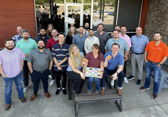 G&G Insurance employees gather for a group photo outside their office, some holding boxes of doughnuts