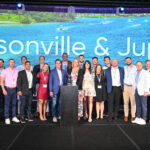 Insurance Office of America employees stand together on a stage in front of a screen reading "Jacksonville & Jupiter"