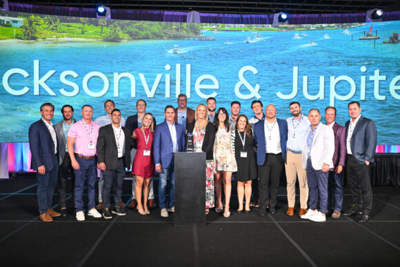 Insurance Office of America employees stand together on a stage in front of a screen reading "Jacksonville & Jupiter"