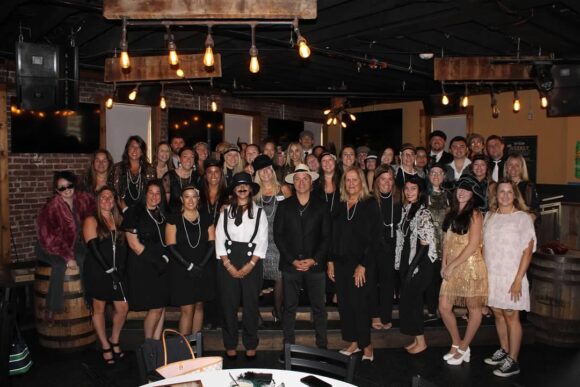 Mackoul Risk Solutions employees, many in 1920s-style costumes, pose for a group photo at an indoor murder mystery lunch
