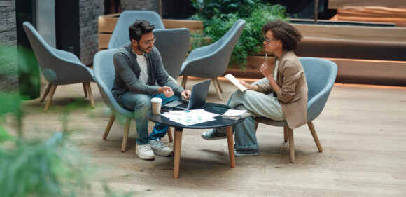 Two colleagues talk over a laptop and coffee while sitting in lounge chairs in a modern office