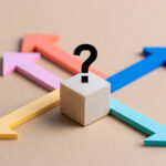 A wooden block with a question mark at a crossroads of colorful arrows pointing in different directions